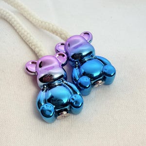Puede incluir: Dos terminales de cordón metálicos con forma de oso que presentan un acabado degradado vibrante, pasando del morado en las orejas a un azul intenso en la base. Cada oso está unido a un cordón grueso trenzado de color blanco roto.