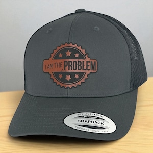 May include: A charcoal gray snapback trucker hat with a mesh back. The front features a distressed copper-colored patch with the text "I AM THE PROBLEM" inside a star-studded border. The hat has a curved bill and a white tag that reads "SNAPBACK".