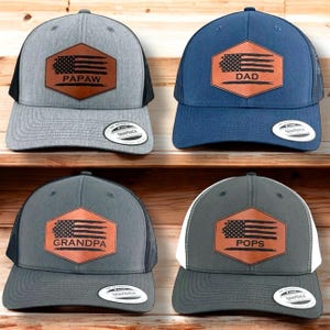 May include: Four trucker hats with a brown leather patch featuring an American flag and a word. The hats are gray, dark gray, navy blue, and gray with a white mesh back. The words on the patches are "Papaw", "Dad", "Grandpa", and "Pops".