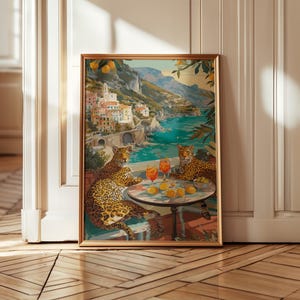 May include: A framed art print depicting two leopards sitting at a round table on a terrace overlooking a coastal town. The table holds two orange cocktails and several yellow lemons. The background features a Mediterranean-style seaside village.