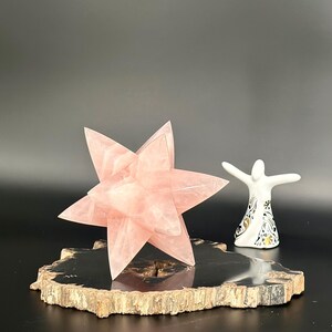 May include: A large, pink, star-shaped crystal sculpture sits on a dark, natural wood slice. A small white figurine with outstretched arms and gold and black floral details stands nearby. The background is black.