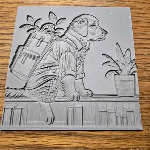 Könnte beinhalten: Eine graue 3D-gedruckte Skulptur eines Hundes, der einen Rucksack trägt und auf einem Regal mit Büchern steht. Der Hund schaut nach rechts und hat einen ernsten Ausdruck. Die Skulptur steht auf einer Holzoberfläche.