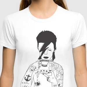May include: A white short-sleeved t-shirt featuring a black graphic illustration of a person with a lightning bolt over one eye, holding a teacup and saucer. The figure has visible tattoos on the arms and chest, including an anchor and roses.