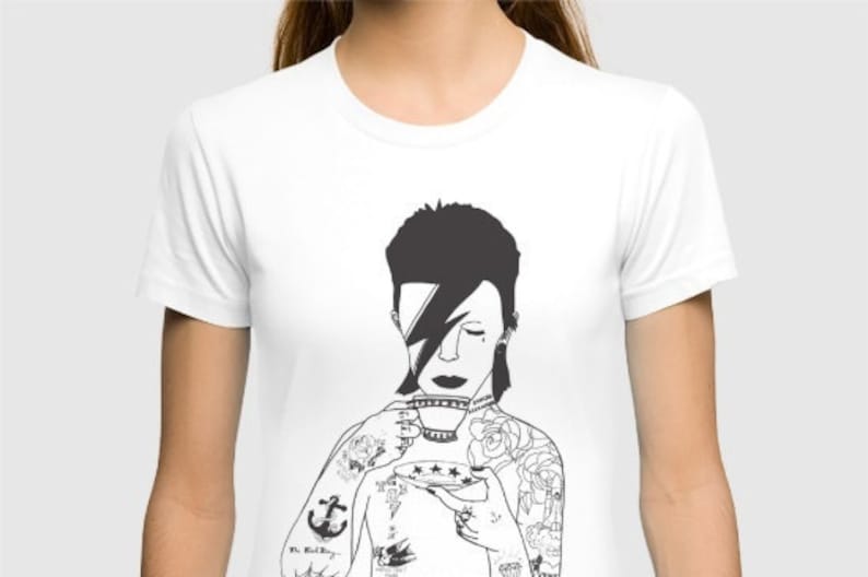 May include: A white short-sleeved t-shirt featuring a black graphic illustration of a person with a lightning bolt over one eye, holding a teacup and saucer. The figure has visible tattoos on the arms and chest, including an anchor and roses.