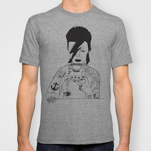 May include: A heather gray t-shirt featuring a black line art illustration of a person with a lightning bolt over one eye, holding a teacup and saucer. The figure has multiple tattoos on their arms and chest, including an anchor and roses.