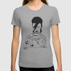 May include: A heather gray t-shirt featuring a black line art illustration of a person with a lightning bolt across their eye, holding a teacup and saucer. The figure has multiple tattoos on their arms and chest, including an anchor and roses.