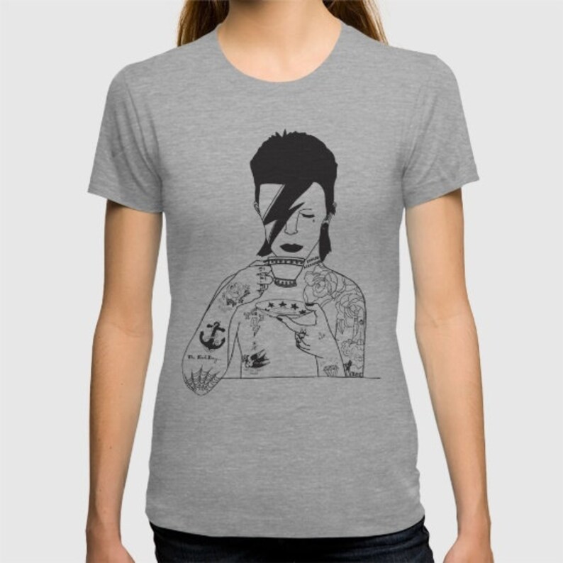 May include: A heather gray t-shirt featuring a black line art illustration of a person with a lightning bolt across their eye, holding a teacup and saucer. The figure has multiple tattoos on their arms and chest, including an anchor and roses.