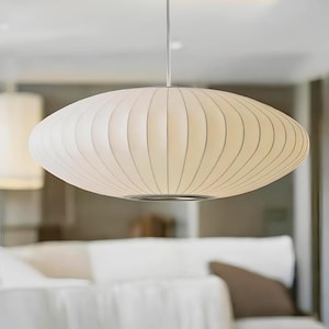 May include: A saucer-shaped pendant light fitting featuring a white, ribbed, translucent material stretched over a frame. The light is suspended by a thin cord and includes a circular metal trim at the bottom centre, emitting a soft glow.