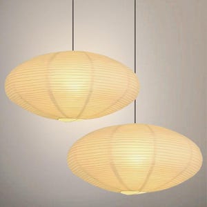 Op de afbeelding: Twee hanglampen met een ovaal, lantaarnachtig ontwerp. De lampenkappen zijn warm off-white van kleur en hebben een geribbelde textuur. De lampen hangen aan zwarte snoeren tegen een neutrale achtergrond.