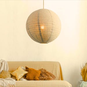 May include: A spherical beige paper lantern hangs from the ceiling, emitting a warm light. Below, a beige sofa is decorated with gold and brown cushions, creating a comfortable and inviting living area.