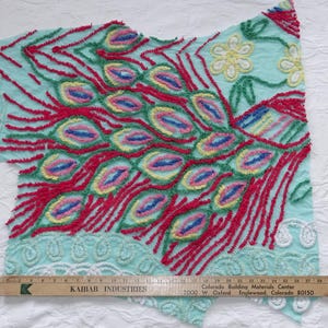 May include: A section of fabric with a peacock feather design. The design features red, green, yellow, and blue accents on a light blue background. The fabric has a textured, embroidered appearance. A wooden ruler is placed across the bottom.