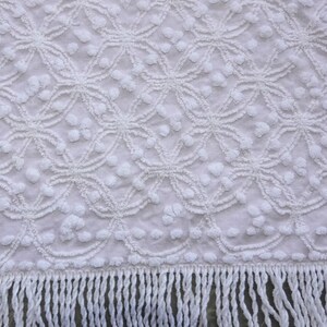 May include: A white fabric textile featuring a raised tufted geometric pattern of interconnected circles and small clusters of dots. The bottom edge of the fabric is finished with a long, straight fringe made of matching white twisted yarn strands.