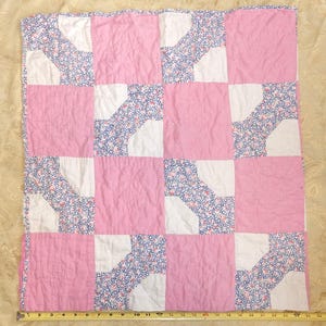 May include: A patchwork quilt with a grid pattern. The quilt features alternating squares of solid pink and white fabric with a floral pattern in blue and white. The quilt is approximately 40 inches wide.