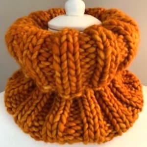 May include: A chunky, hand-knitted cowl in a warm, golden-orange hue. The thick yarn creates a textured, ribbed pattern, perfect for cold weather. The cowl is displayed on a white form, showcasing its circular design and cozy appeal.