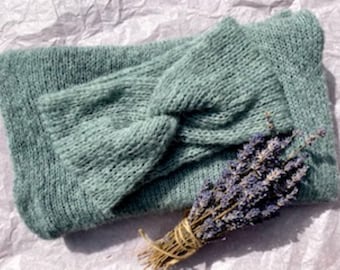 May include: A sage green knitted headband featuring a central twist detail, paired with a matching sage green knitted scarf. A small bundle of dried purple lavender tied with twine is placed on top of the folded knitted items.