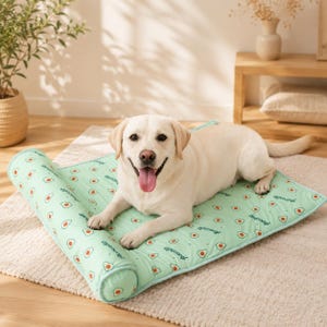 May include: A light green rectangular pet bed featuring a repeating pattern of small avocado halves and the word Avocado in cursive script. The bed has a cylindrical bolster along one side and a flat cushioned base for resting.