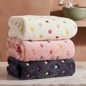 May include: A stack of three folded plush towels with a polka dot pattern. The top towel is cream with multicolored dots, the middle towel is light pink with multicolored dots, and the bottom towel is dark charcoal gray with multicolored dots.