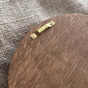 May include: Close-up of a wooden, circular object with a brass sawtooth hanger attached. The wood has a rich, brown color and visible grain. The hanger is centered and secured with small, gold-colored brackets.