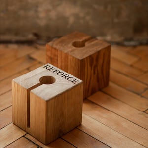 Op de afbeelding: Twee houten blokken met een sleuf en een cirkelvormige opening. Op het voorste blok staat het woord "REFORCE" gegraveerd. De blokken zijn gemaakt van lichtbruin hout en staan op een houten vloer.