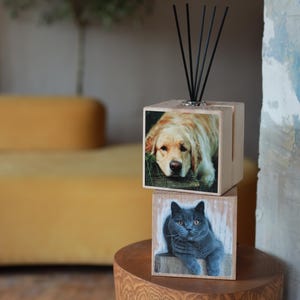 May include: A wooden essential oil diffuser with a photo of a golden retriever on the top cube and a gray cat on the bottom cube. The diffuser has black reeds and sits on a wooden surface.