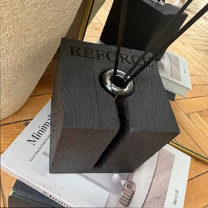 Puede incluir: Un difusor de madera negra con la palabra "REFORCE" grabada en la parte delantera. El difusor tiene un anillo plateado alrededor de la abertura para las cañas y una ranura vertical. Las cañas negras se insertan en el difusor. El difusor se encuentra encima de una pila de libros.