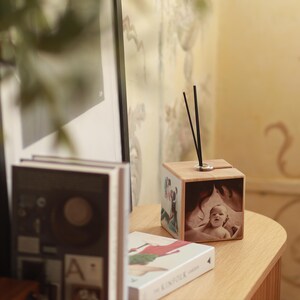 Könnte beinhalten: Ein Würfel-Diffusor aus Holz mit einem silbernen Ring und schwarzen Stäbchen. Der Würfel zeigt gedruckte Bilder, darunter ein Babyporträt. Er steht auf einem Holztisch neben Büchern.