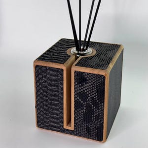 May include: A decorative reed diffuser with a square wooden frame and black faux snakeskin panels. The diffuser has a clear glass insert holding black reeds. The wooden frame has a natural wood finish.