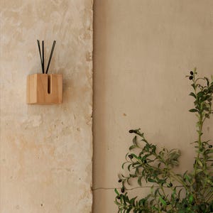 May include: A wooden essential oil diffuser with black reeds. The cube-shaped diffuser is mounted on a textured, light-coloured wall. The background features a plant with green leaves and small, dark berries.