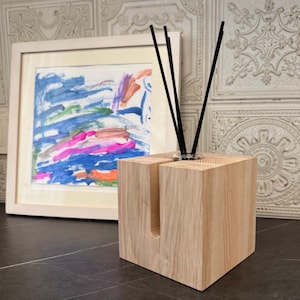 Könnte beinhalten: Ein hölzerner Diffusor für ätherische Öle mit schwarzen Stäbchen. Der quadratische Block hat einen Schlitz in der Vorderseite. Ein gerahmtes abstraktes Gemälde mit blauen, rosa und orangen Farbtönen befindet sich im Hintergrund. Der Diffusor steht auf einer dunklen Oberfläche.