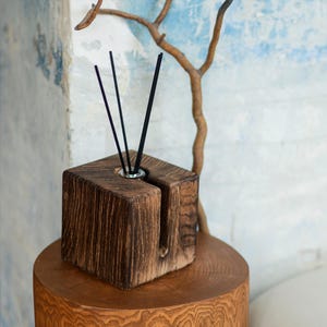 Può includere: Un diffusore di oli essenziali quadrato in legno marrone scuro con bastoncini neri. Poggia su un tavolino rotondo in legno. Lo sfondo presenta un muro strutturato e un ramo decorativo.