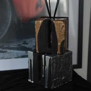Puede incluir: Un difusor decorativo de dos niveles. El cubo superior es negro con pintura dorada goteando, y el cubo inferior es negro con pintura plateada goteando. Las cañas negras se insertan en el cubo superior.