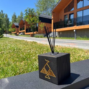 Op de afbeelding: Een zwarte, kubusvormige etherische olie diffuser met een gouden geometrisch vogelontwerp. De diffuser heeft zwarte rietjes en staat op een donkergrijze ondergrond. De achtergrond toont een grasveld en houten gebouwen.