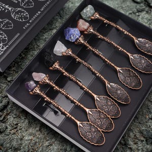 May include: Set of six ornate copper spoons with different coloured gemstone tops. The spoons have a floral design and the text "For Modern Witches" is visible on the box.
