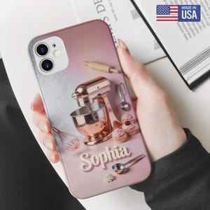 May include: A pink and silver phone case featuring a rose gold stand mixer, cupcakes, and baking tools. The name "Sophia" is displayed in white script. The case has a "Made in USA" sticker.