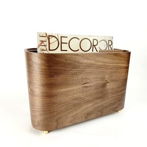 May include: A wooden magazine rack with a curved design and a warm brown finish. The rack has a rectangular shape with rounded edges and sits on small, gold-coloured feet. A magazine with the word "DECOR" is visible inside the rack.