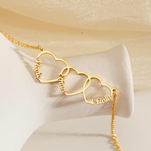 May include: Gold-colored necklace featuring three interconnected heart pendants. Each heart is inscribed with a name: Molly, Olivia, and Olivia. The necklace is displayed on a white surface against a soft, neutral background.