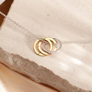 Puede incluir: Un collar de cadena plateada con tres colgantes circulares entrelazados en tonos oro, oro rosa y plata. Cada anillo está grabado con un nombre, incluyendo el texto visible ETHAN, ARIA y LUCAS, suspendido de una delicada cadena de eslabones.