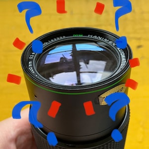 Puede incluir: Lente de cámara automática Hanimex negra con detalles en verde y dorado. La lente tiene un diámetro de 52 mm y el texto "Hanimex Automatic" es visible. La lente se sostiene sobre un fondo amarillo con formas abstractas azules y rojas.
