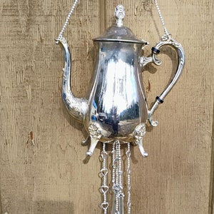 Puede incluir: Carillón de tetera de plata con una cadena de corazones y cinco cucharas de plata colgando de él. El carillón está colgado de una superficie de madera.