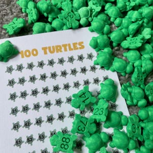 Op de afbeelding: Een witte kaart met de tekst "100 TURTLES" is omringd door talrijke kleine, groene schildpadvormige spelstukken. Elke schildpad heeft een nummer op zijn rug en de kaart heeft genummerde vakjes.