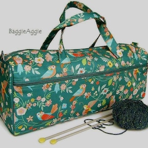May include: A teal fabric duffel bag with a floral and bird pattern, featuring the text BaggieAggie. The bag has two top handles and a zip closure. Beside the bag sits a ball of dark green yarn and two long knitting needles with yellow tips.