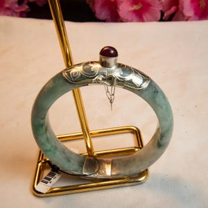 May include: A jade bangle bracelet with silver accents and a small red gemstone. The bracelet is displayed on a gold-colored stand. The jade is a pale green color with darker green marbling.