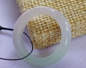 May include: A translucent jade bangle bracelet with a pale green hue. The circular bracelet is displayed against a textured beige background, with a black cord attached. The jade has a smooth, polished surface.