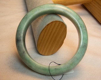 Op de afbeelding: Een lichtgroene jade armband. De gladde, gepolijste armband heeft een cirkelvorm en een subtiel gemarmerd patroon. De jade is een zachte, gedempte groene kleur met donkerdere groene en grijze insluitsels. De armband wordt op een wit oppervlak gepresenteerd.