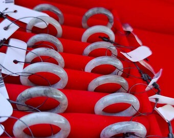 May include: A display of jade bracelets with gray and white tones, arranged on a red velvet surface. Each bracelet is round and has a unique pattern. The bracelets are attached to tags with black strings, suggesting they are for sale.