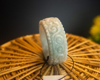 May include: A white jade bangle with a carved design of swirling patterns. The bangle is on a woven surface.