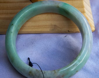 Pode incluir: Uma pulseira de jade com uma superfície lisa e polida. A pulseira apresenta uma mistura de tons verde claro e branco, com variações sutis de cor. O design circular é uma peça de joalheria clássica.