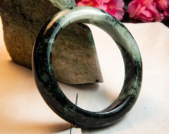 May include: A polished jade bangle bracelet with a dark green and light green marbled pattern. The bracelet is circular and sits on a white surface, with a tag attached. Pink flowers and a green stone are in the background.