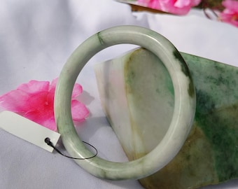Pode incluir: Uma pulseira de jade verde claro. A pulseira tem uma superfície lisa e polida com variações de cor. O jade está sobre um pedaço de jade e uma superfície branca. Flores cor-de-rosa ao fundo.