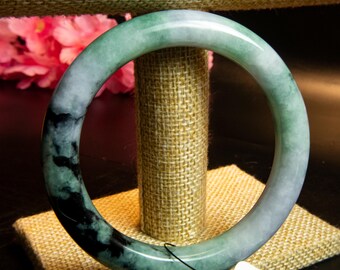 May include: A jade bangle bracelet with a mix of green, white, and black hues. The polished stone is displayed on a textured stand, showcasing its natural color variations. The bracelet is a circular shape, and the colors blend together.